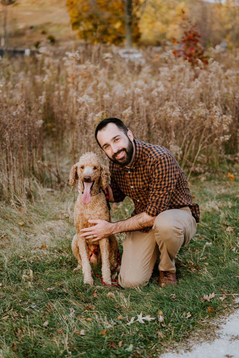 A man kneeling in the grass and smiling next to her brown dog 