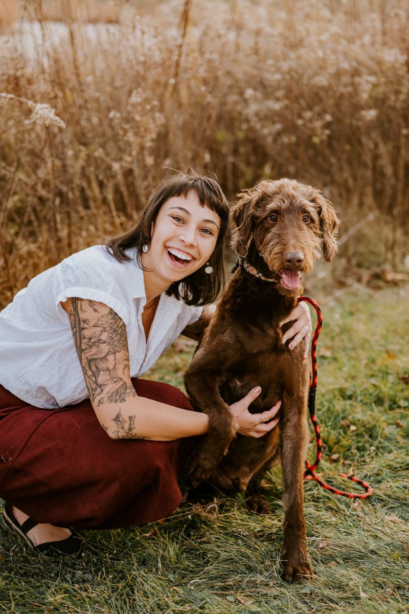 A woman kneeling in the grass smiling and holding onto her brown dog 