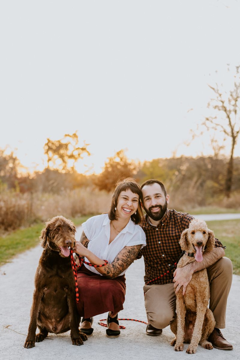 A couple kneeling down and smiling on a paved path holding both of their brown dogs 