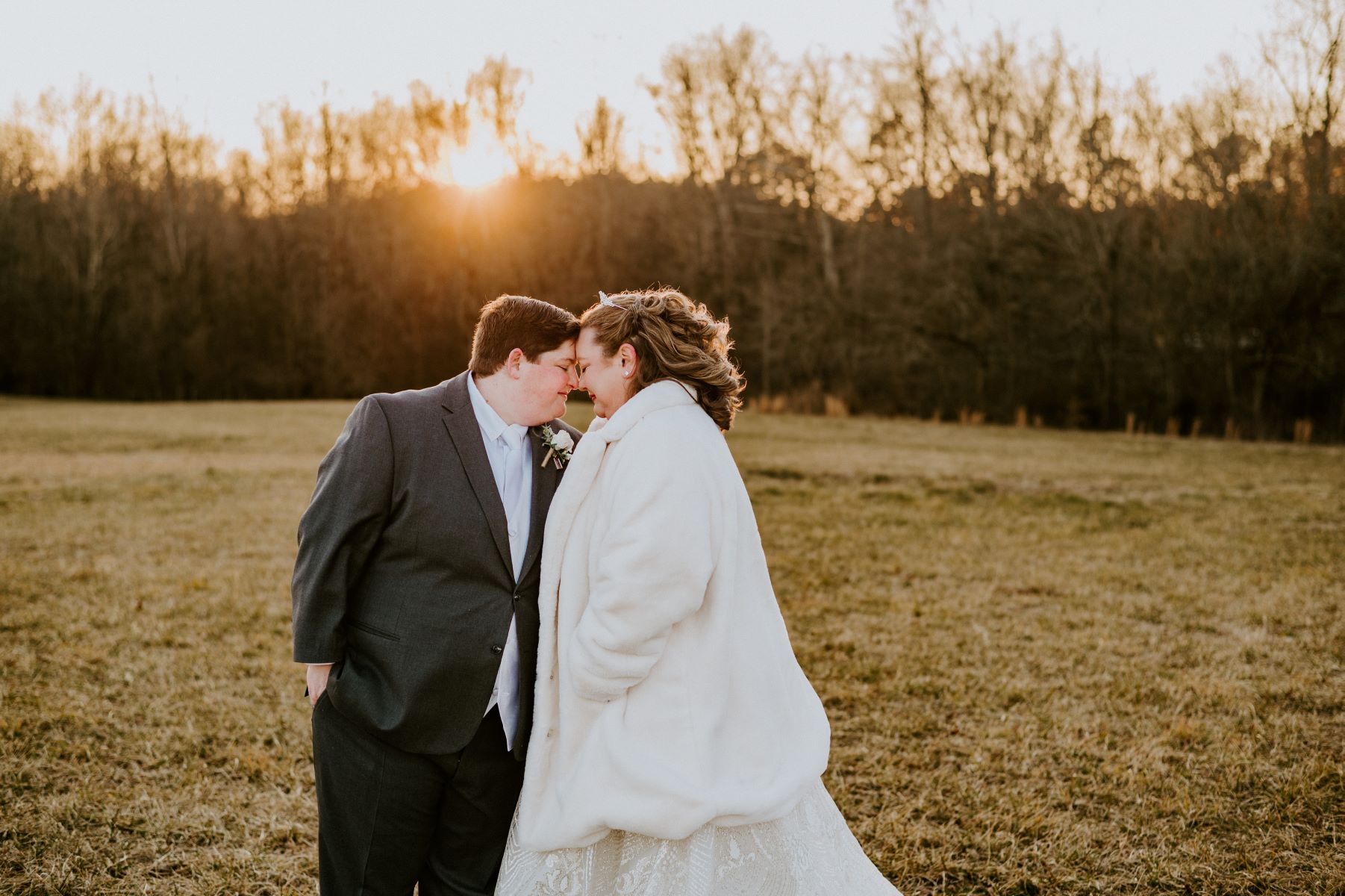 A couple in a field together the sun is setting behind them they have their heads pressed against each other and are smiling one is wearing a suit and the other is wearing a white fur coat and a white wedding dress 