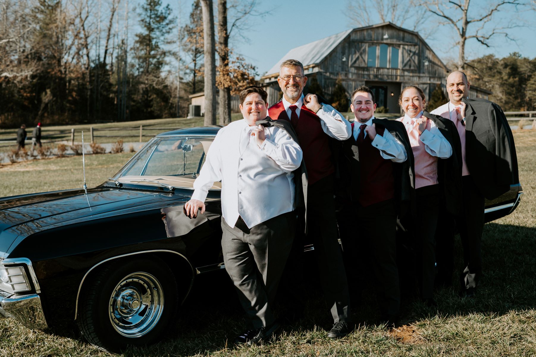 A group standing in front of an old black car with a barn in the background they all have their dress jackets slung over their shoulders 