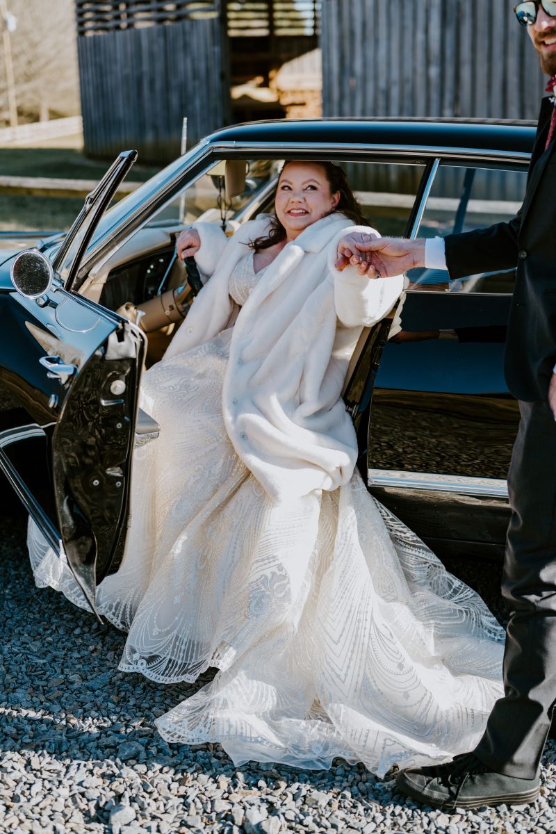 A woman in a car who is wearing a white fur coat and a white wedding dress and someone is helping them get out of the car they are seated in the car door is open 