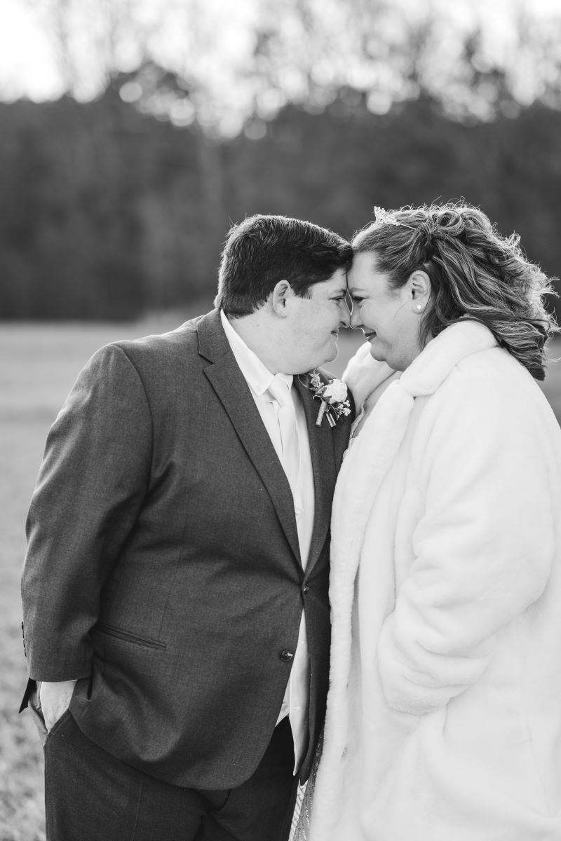 A couple smiling at each other with their heads pressed together one is wearing a white fur coat the other is wearing a suit 