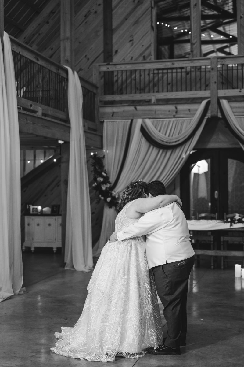 A couple dancing together at their reception with their arms wrapped around each other one is wearing a white wedding dress the other is wearing a white vest and black pants 