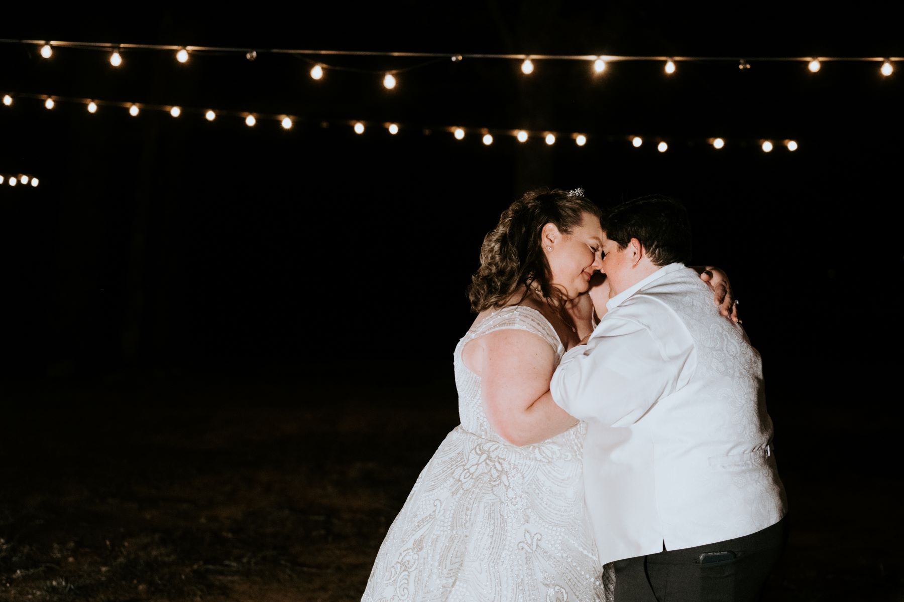 A couple dancing together at their wedding reception underneath string lights one is wearing a white wedding dress and the other is wearing a white vest and white button up and black pants 