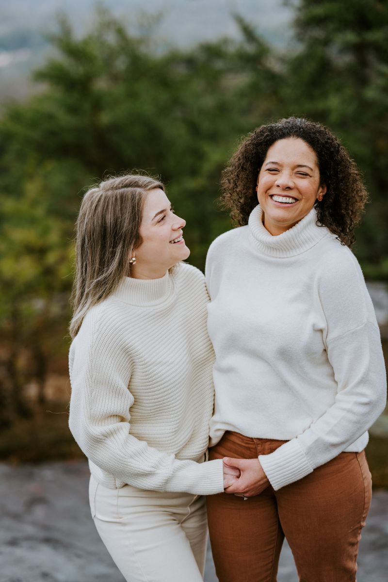 Two women laughing and smiling one woman is wearing a white shirt and white pants the other woman is wearing a white sweater and brown pants 