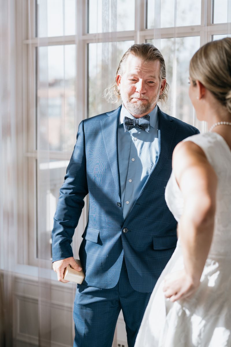 A woman in a white wedding dress doing a first look with her father who is wearing a blue suit 