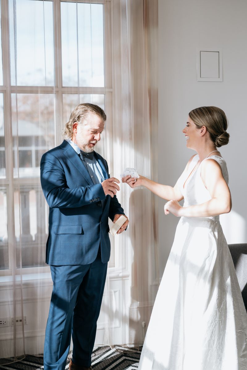 A woman in a white wedding dress offering her father a tissue her father is wearing a blue suit 