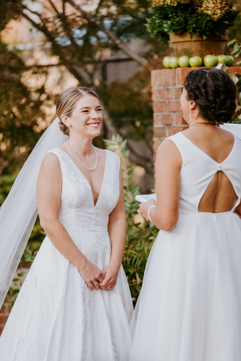 Two women at their wedding ceremony they are both wearing white dresses and one woman is reading vows to her partner and her partner is smiling  