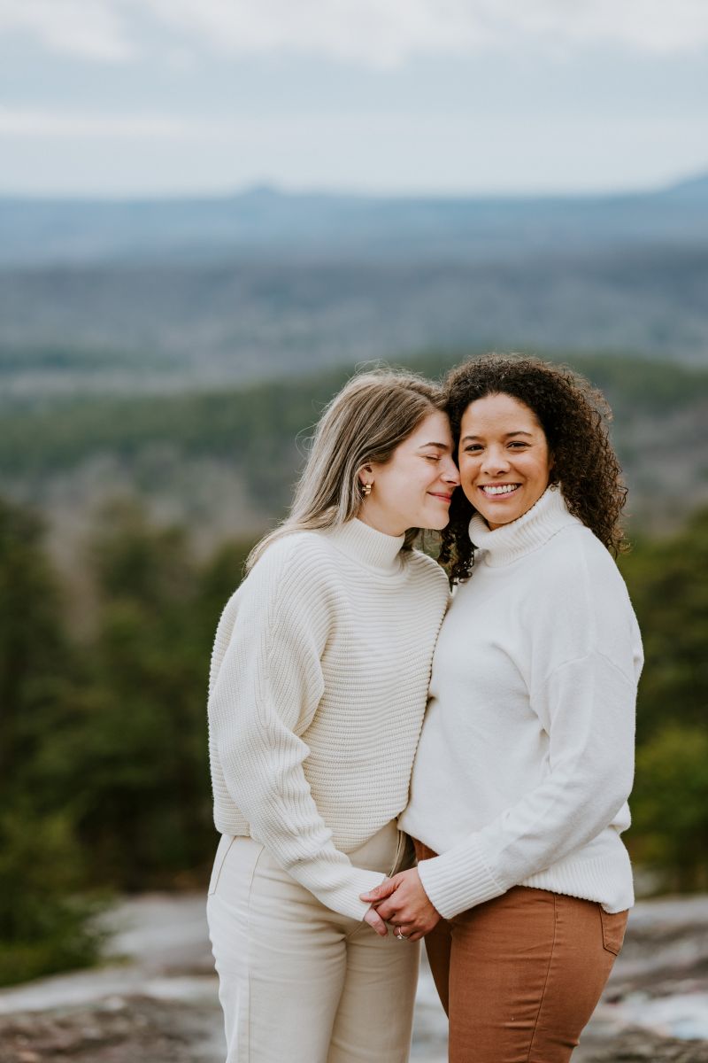 Two women holding hands one woman has her head pressed against the side of her partner's head and is smiling the other woman is looking foward andn smiling both women are wearing white sweaters 