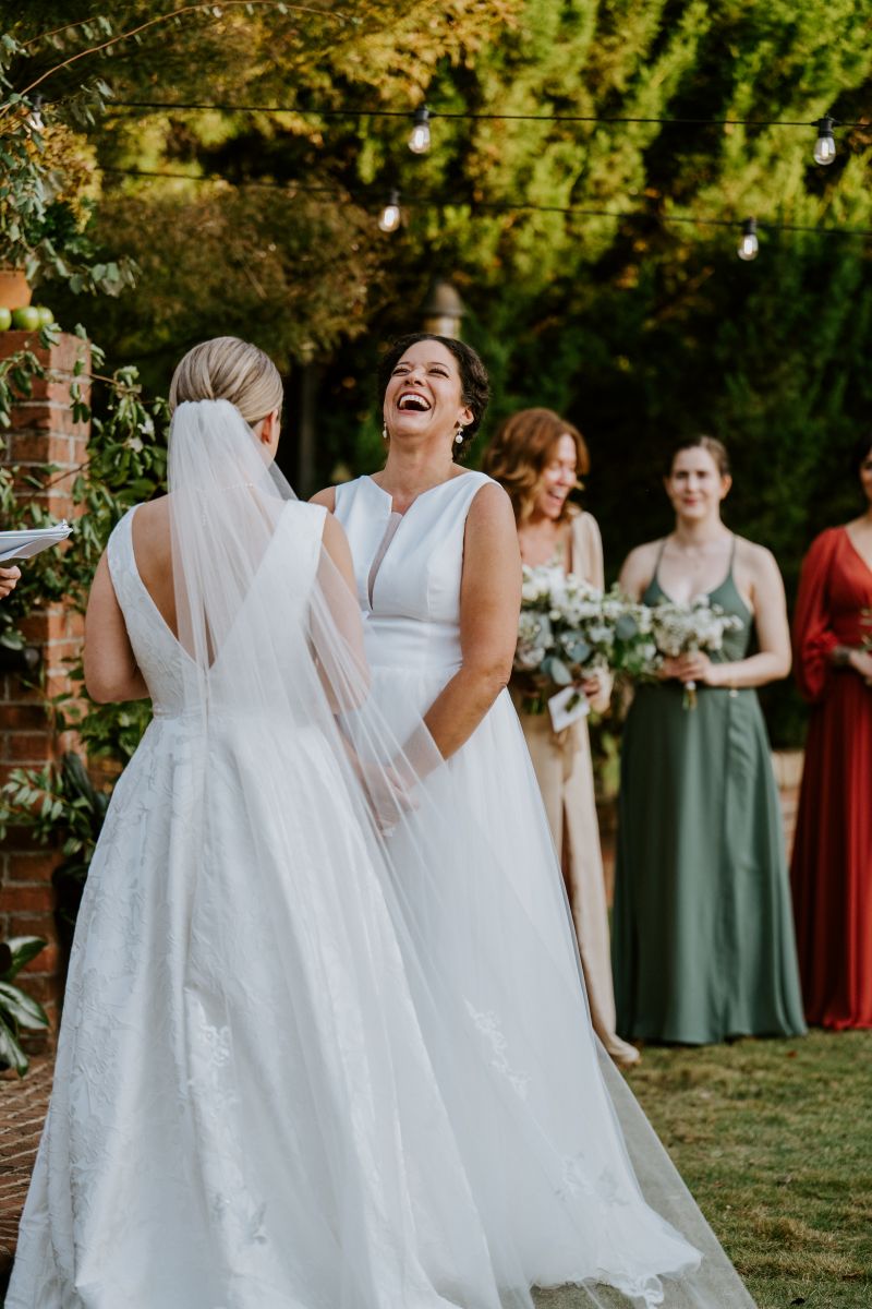 Two women at their wedding ceremony they are both wearing white dresses and one woman is reading vows to her partner the other partner is laughing 