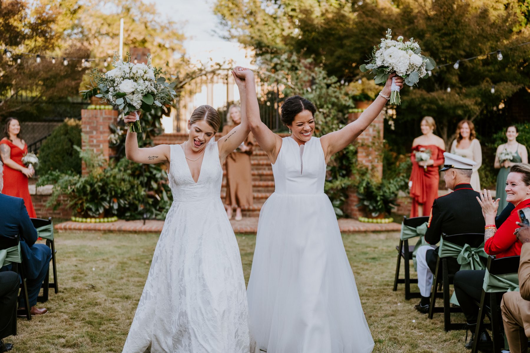 Two women celebrating their marriage at an outdoor ceremony both women are holding their white wedding bouquets up in the air and their wedding guests are sitting and cheering for them 