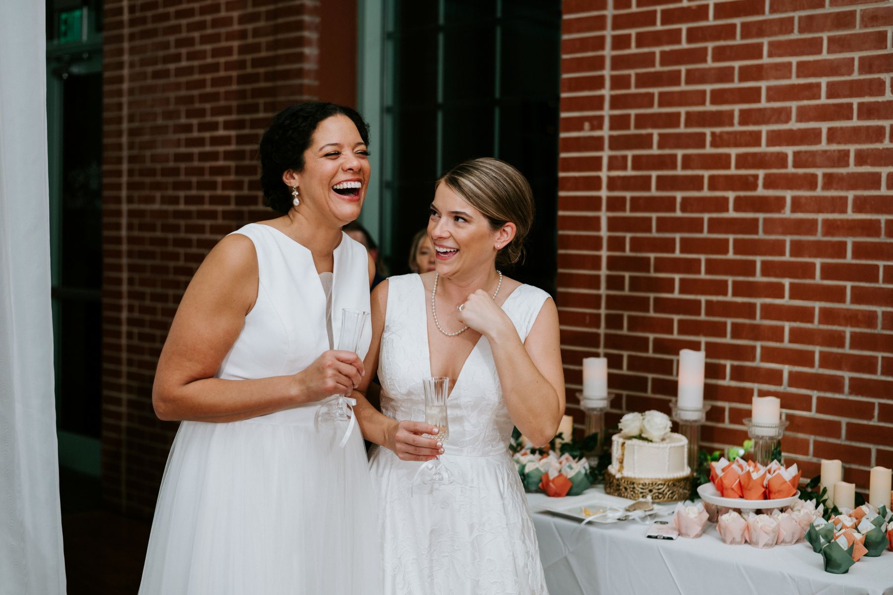 Two women holding champagne glasses and laughing with each other at their wedding reception behind them is their weding cake on a table 