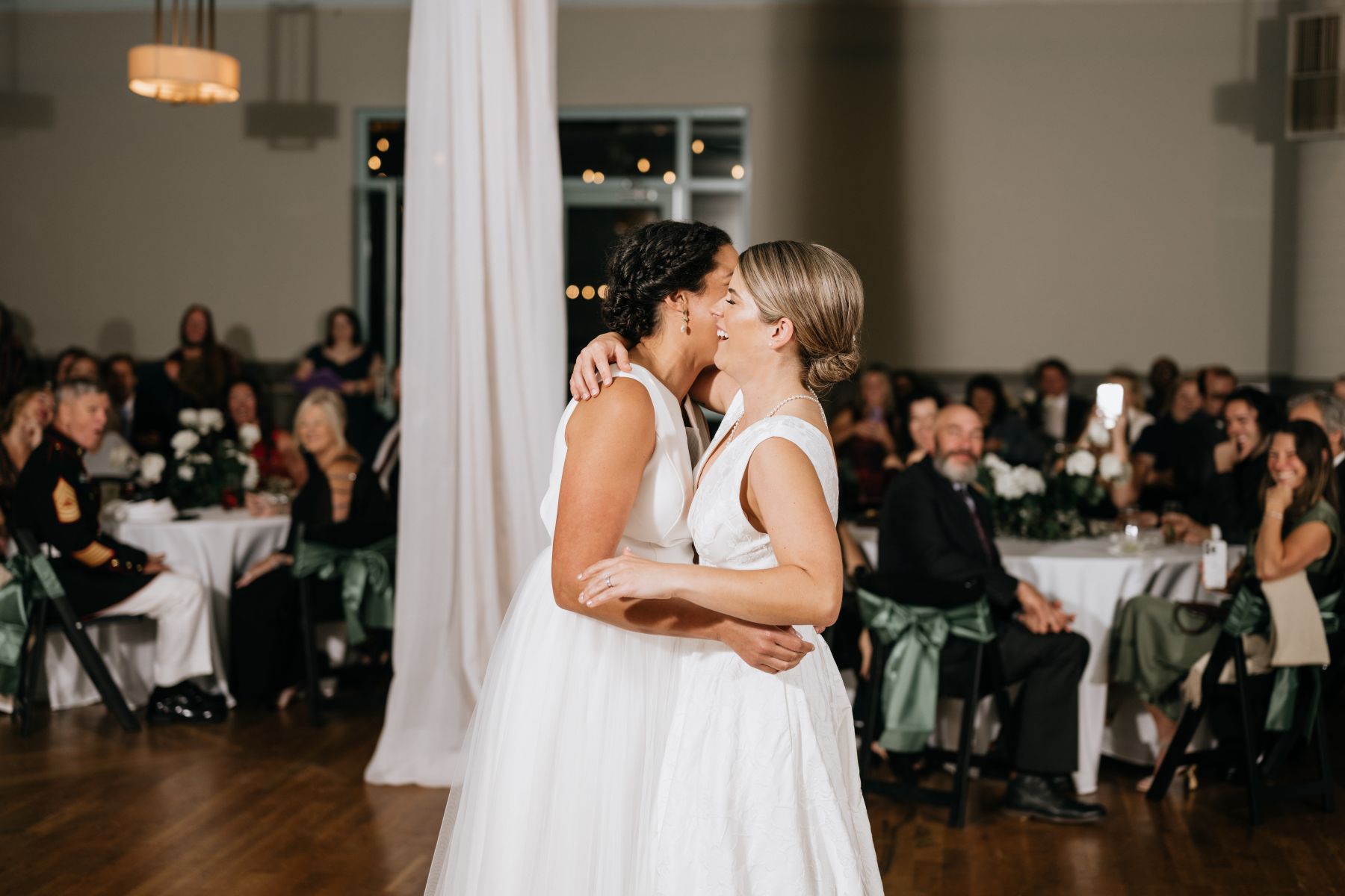 Two women in white wedding dresses at their reception sharing their first dance and laughing with each other and behind them their guests are watching them 