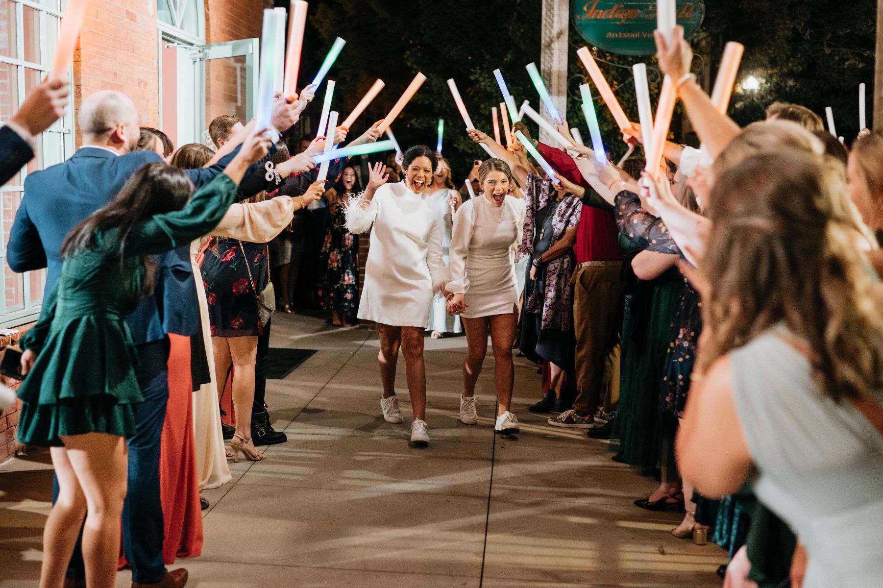 Two women leaving their wedding reception holding hands and their guests are holding colorful light tubes up in the air and cheering for them 