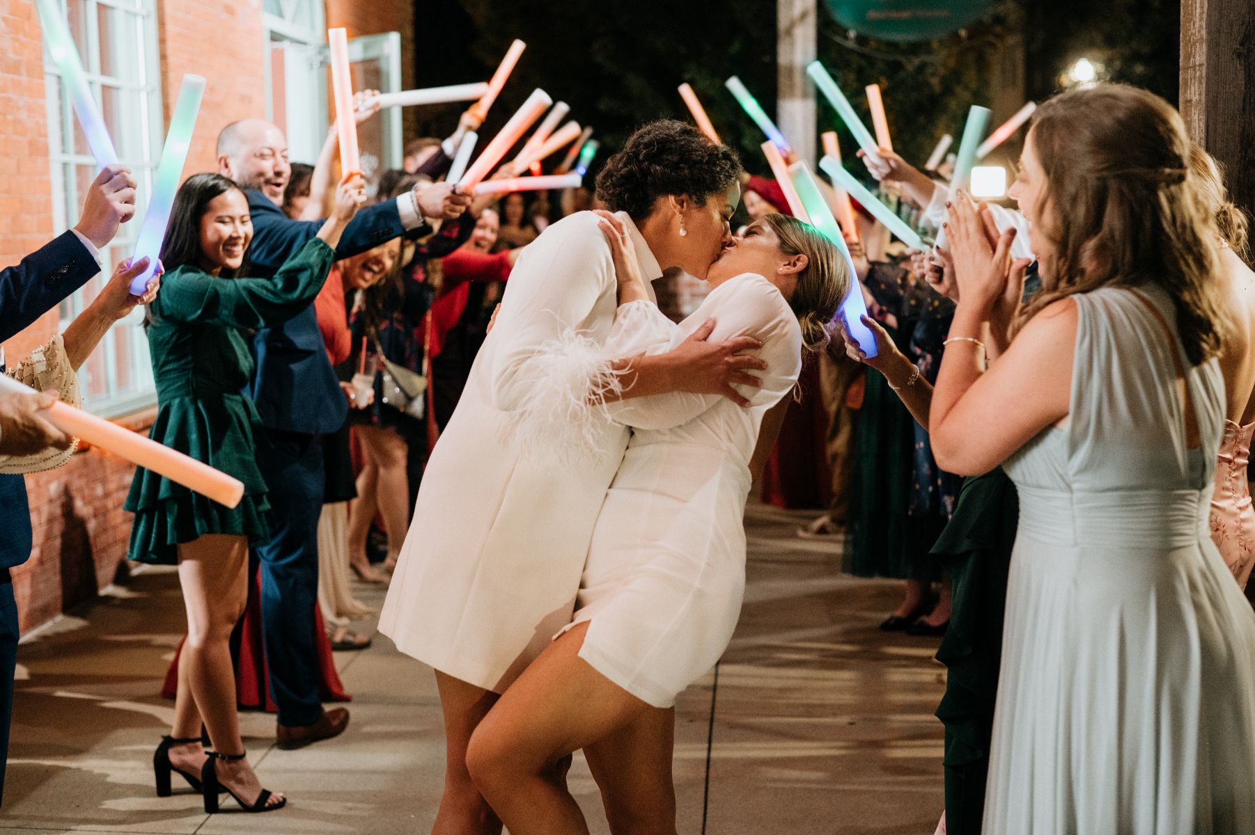 Two women kisisng outside both are wearing short white dresses their guests are behind them cheering and holding colorful light tubes 