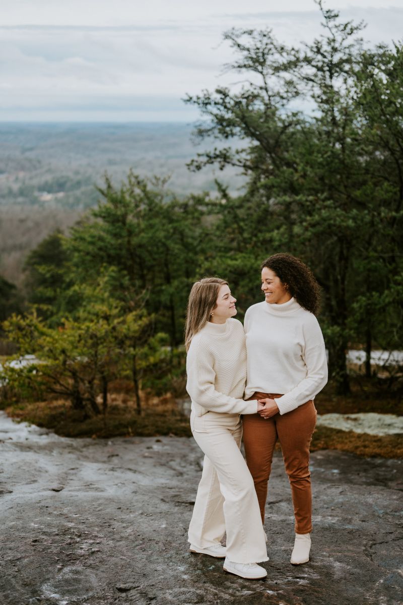 Two women standing on top of a rock that is surrounded by trees and holding hands and smiling at each other both women are wearing white sweaters 