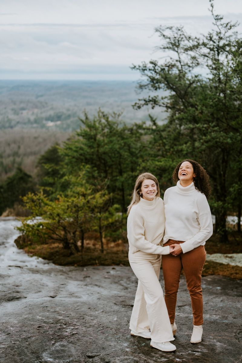 Two women standing on top of a hill surrounded by trees holding hands and laughing with each other both women are wearing a white sweater 