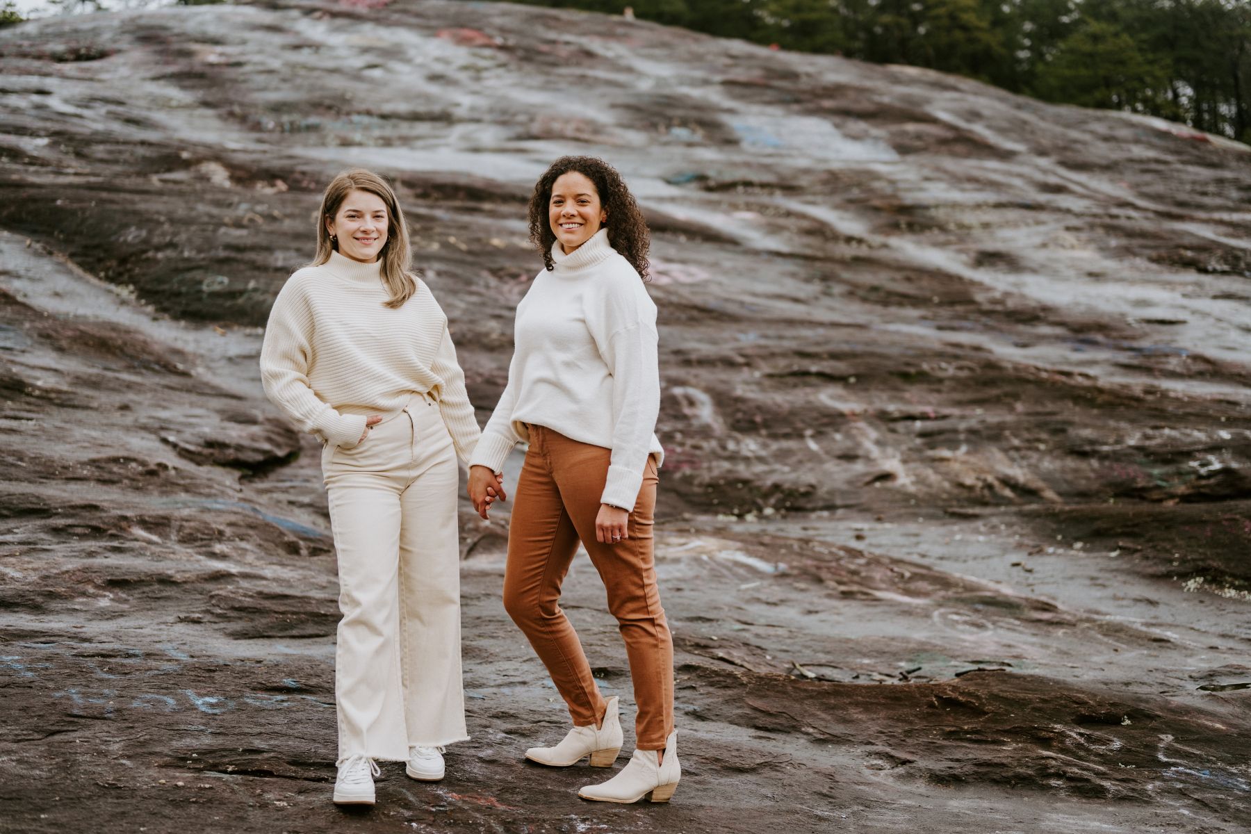 Two women standing on top of a large rock smiling one woman is wearing a white sweater and white pants and has her hand in her pocket with her other hand she is holding her partner's handd her partner is wearing a white sweater and brown pants 