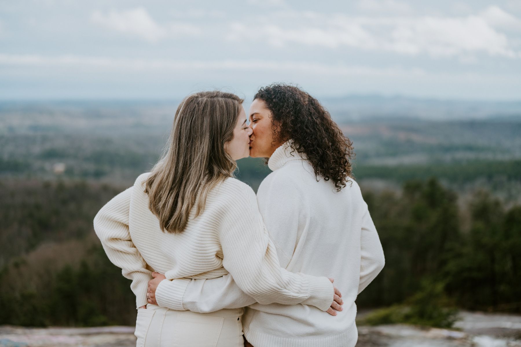 Two women in white sweaters kissing on top of a rock overlooking a valley with their arms around each other 