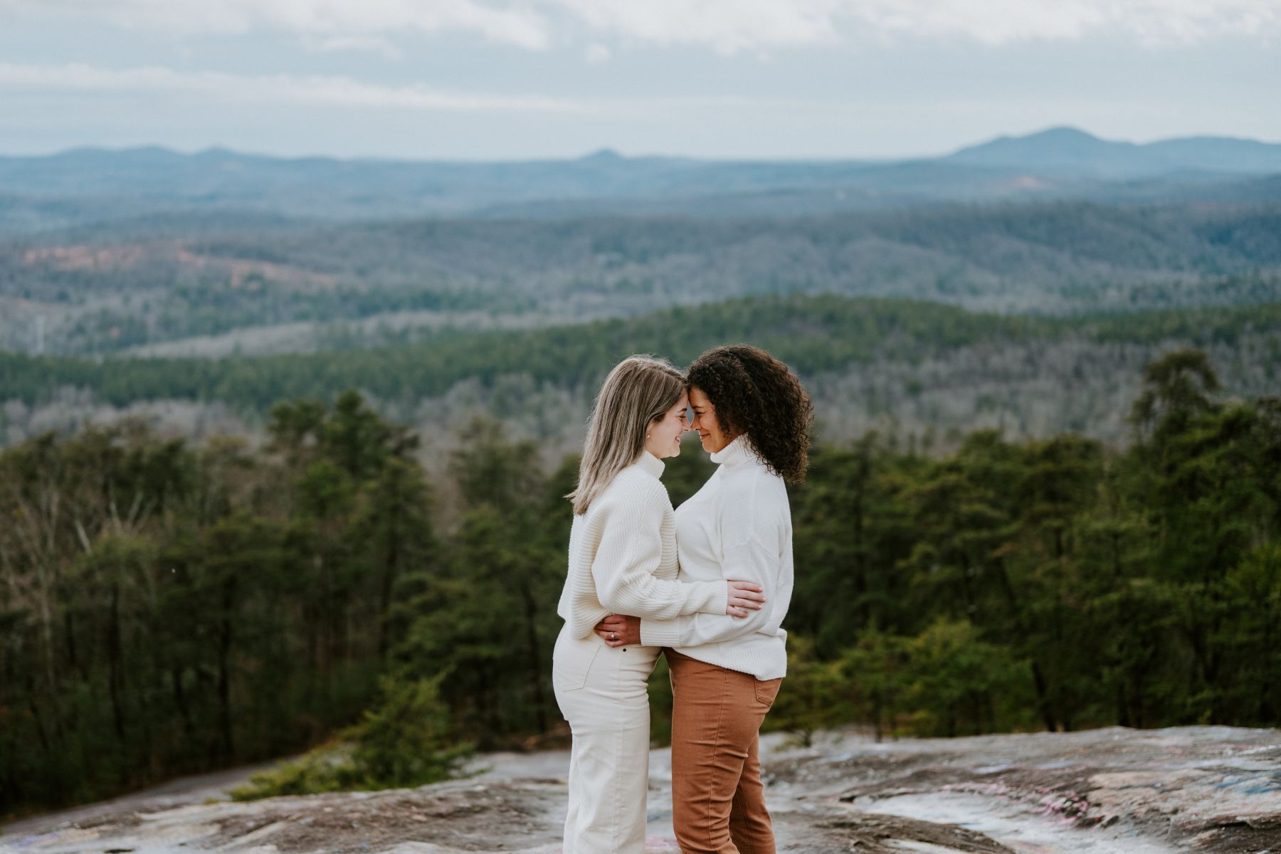 Two women on top of a rock overlooking a valley with their heads pressed against each other and holding one another and smiling both women are waering white sweaters 