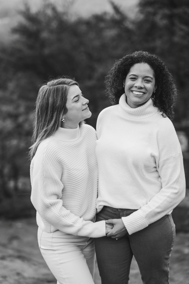 two women holding hands one woman is looking at the other and softly smiling and the other woman is smiling and looking forward 