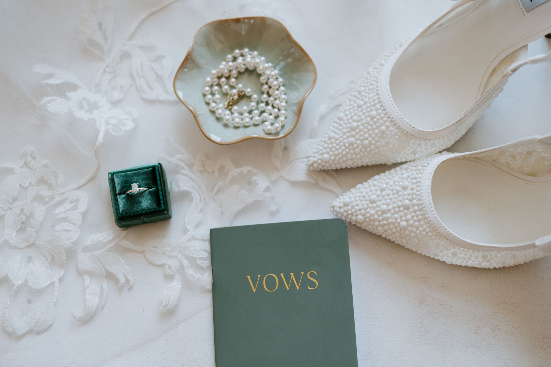 wedding details that include a diamond ring, lace, pearls, heels, adn a vow book 