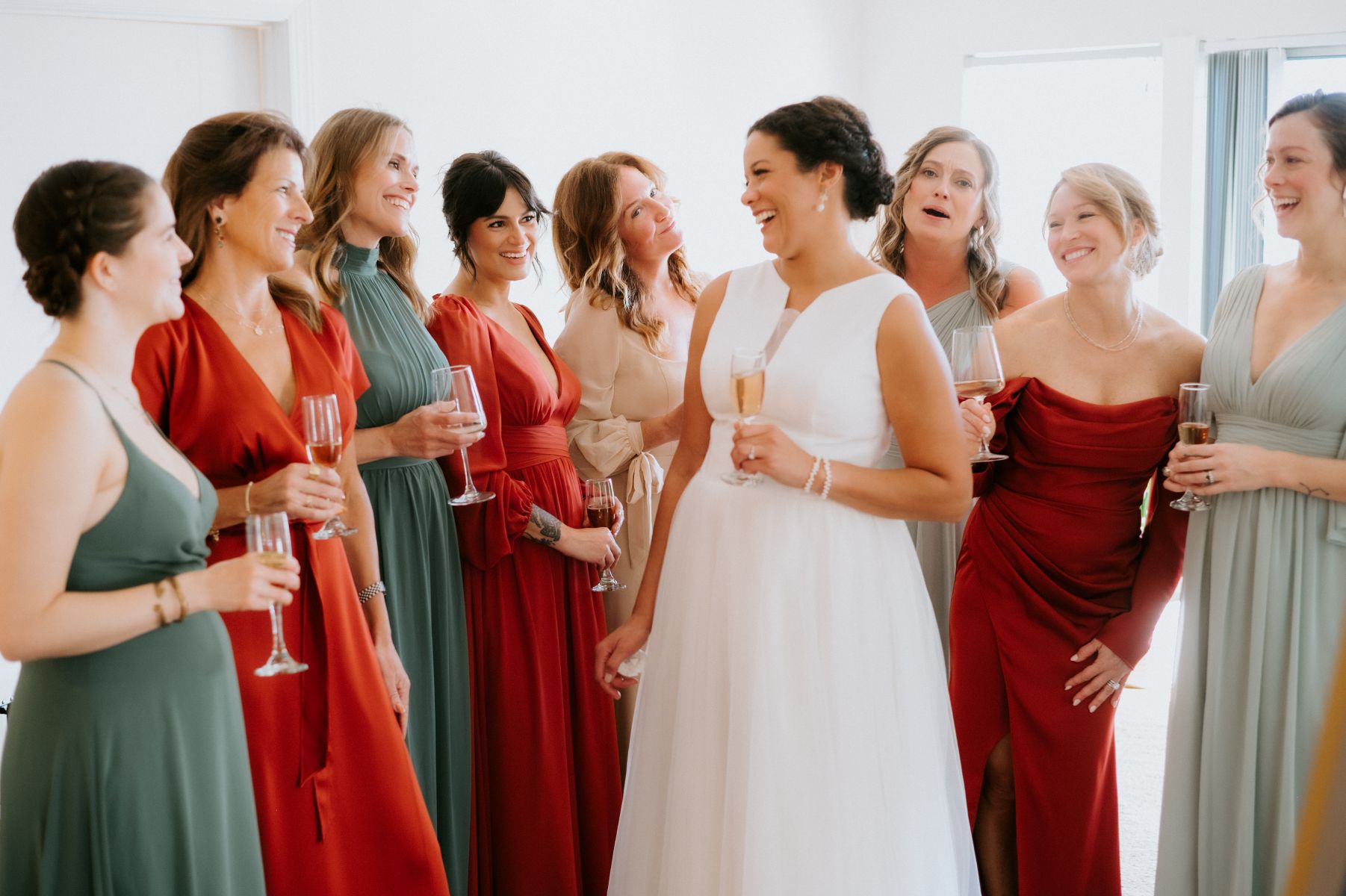 A woman is a white wedding dress holding a glass of champagne and surrounded by her wedding party in a room her  the members of her wedding party are wearing desert color themed dresses