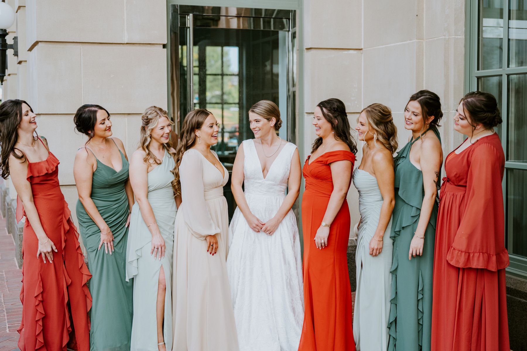 A woman in a white dress standing outside a building with her wedding party made up of all women who are wearing desert colored themed dresses 