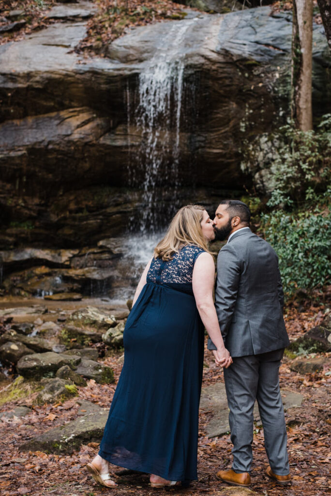 A couple holding hands and kissing next to a waterfall 
