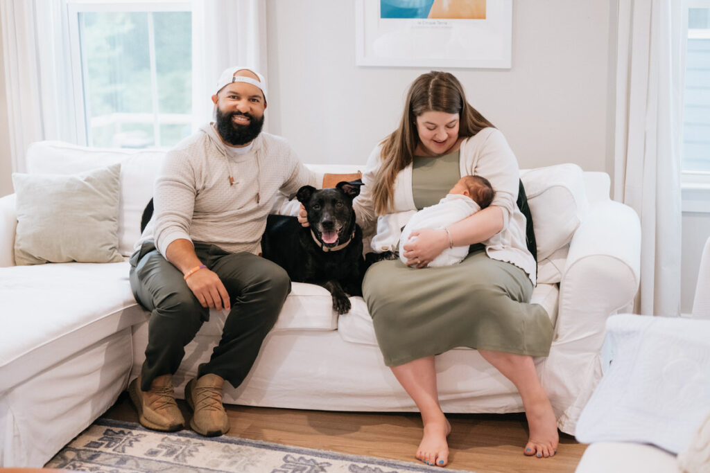 Parents sitting on a couch with a dog in between them as one holds a newborn 
