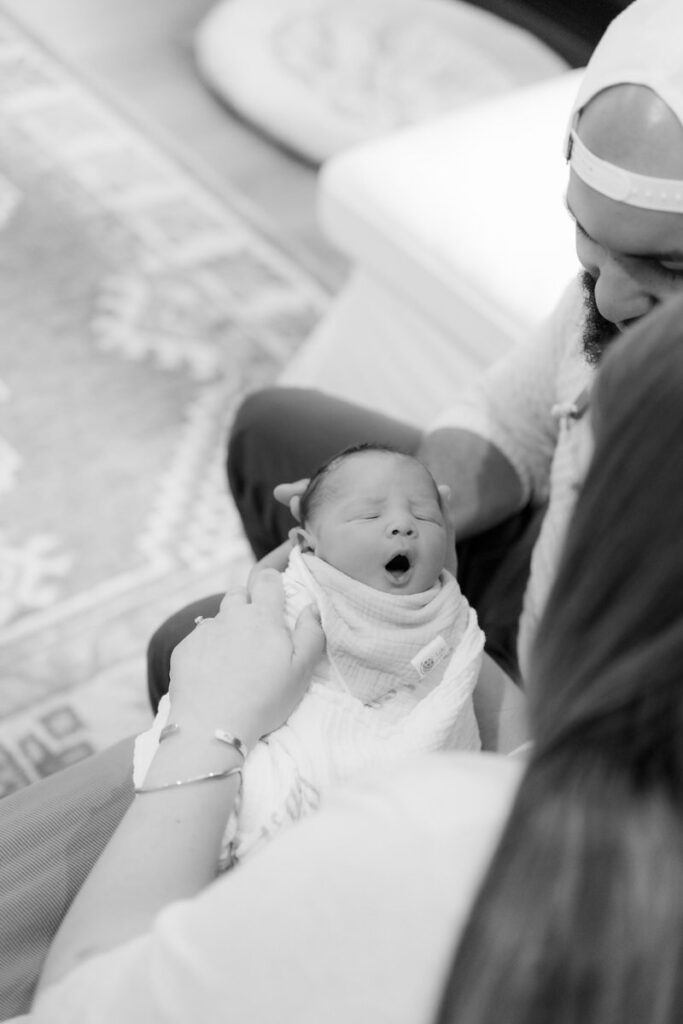 Two parents sitting on a couch holding a yawning newborn in between them 
