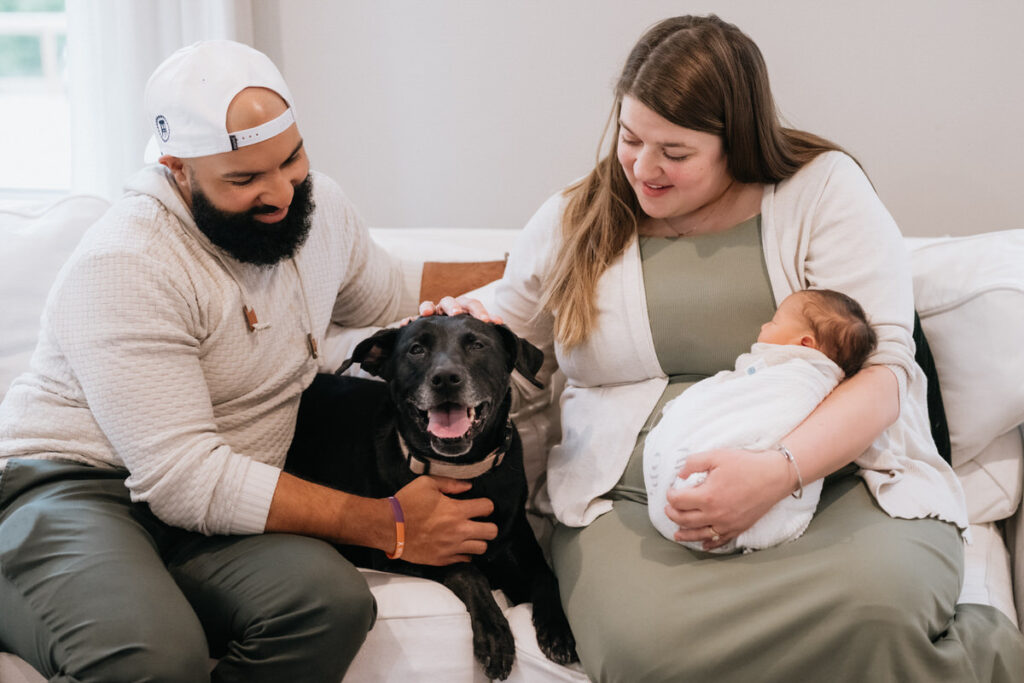 Two parents sitting on a couch petting a dog in between them while one holds a newborn in their lap 
