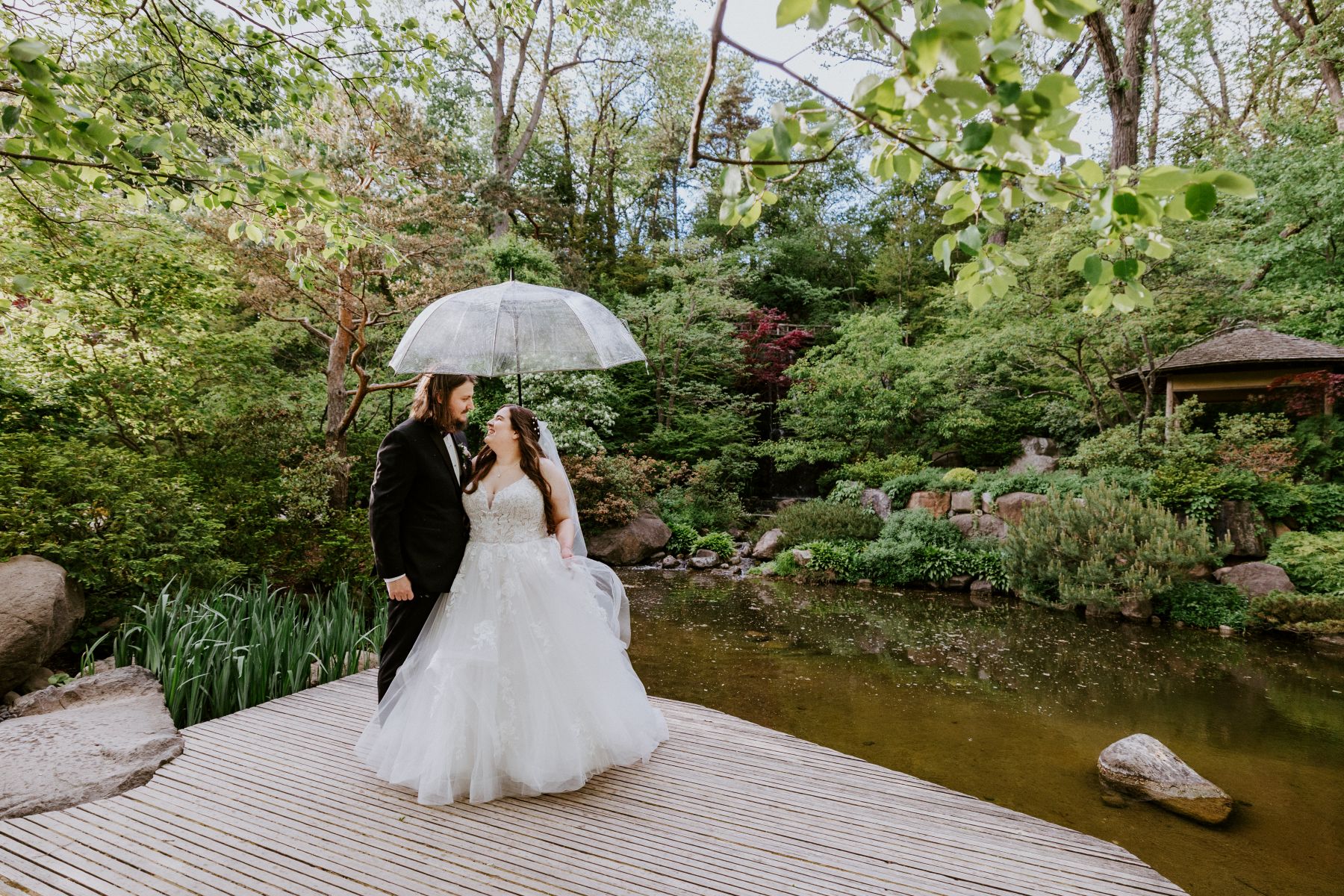 a couple next to a koi pond holding a clear umbrella they are embracing and smiling at each other the woman is wearing a white wedding dress and veil the man is wearing a black suit