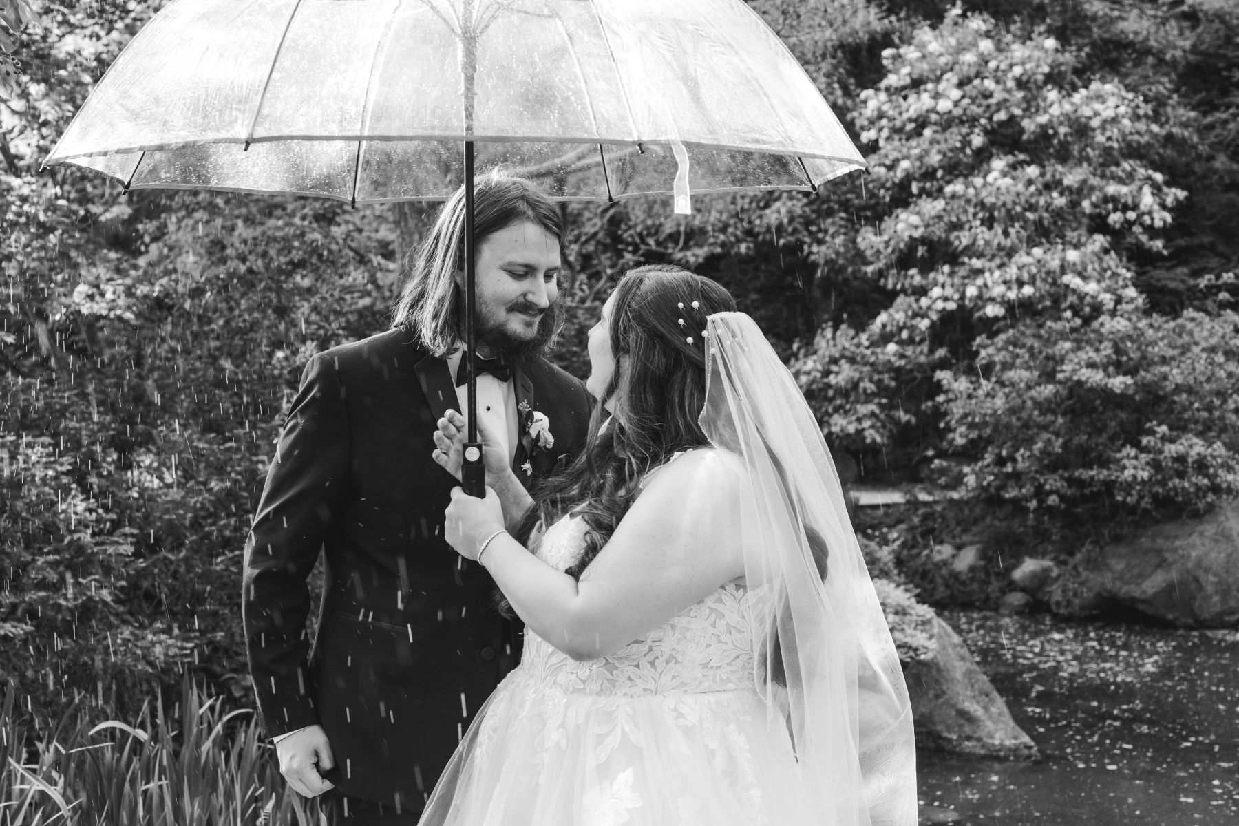 a couple smiling at each other and holding a clear umbrella while it's raining the woman is wearing a white wedding dress and veil and the man is wearing a black suit