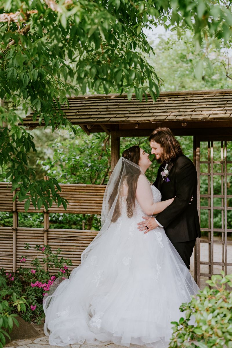 a couple embracing next to a wooden pergola the woman is wearing a white wedding dress and veil the man is wearing a black suit
