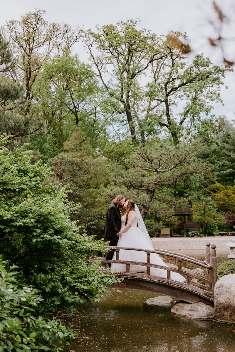 a couple kissing on a wooden bridge in anderson japanese gardens during their wedding at the gardens the woman is wearing a veil and white wedding dress the man is wearing a black suit