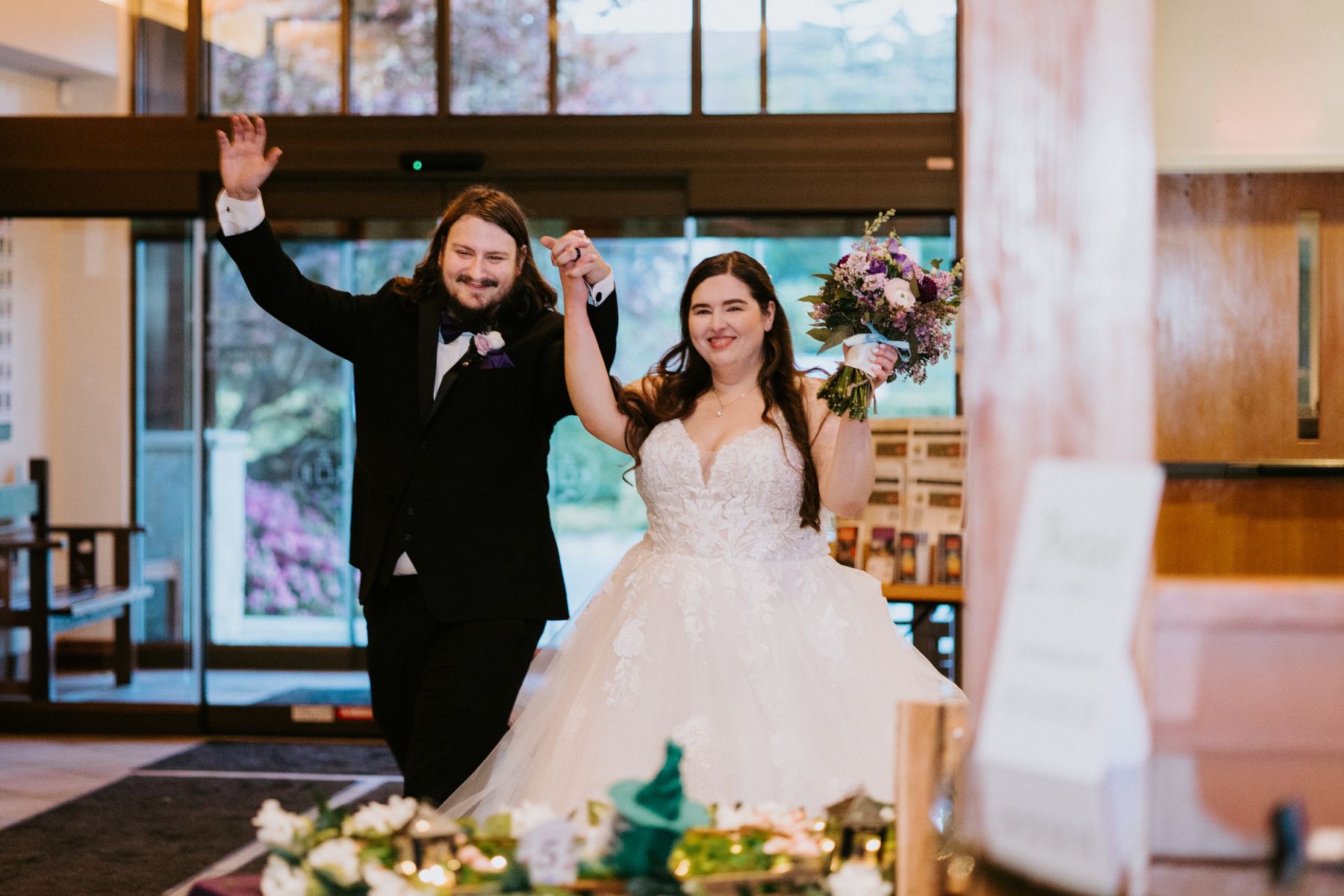 a couple walking into their wedding reception holding their hands in the air and smiling the woman is wearing a white wedding dress and holding a bouquet of flowers the man is wearing a black suit
