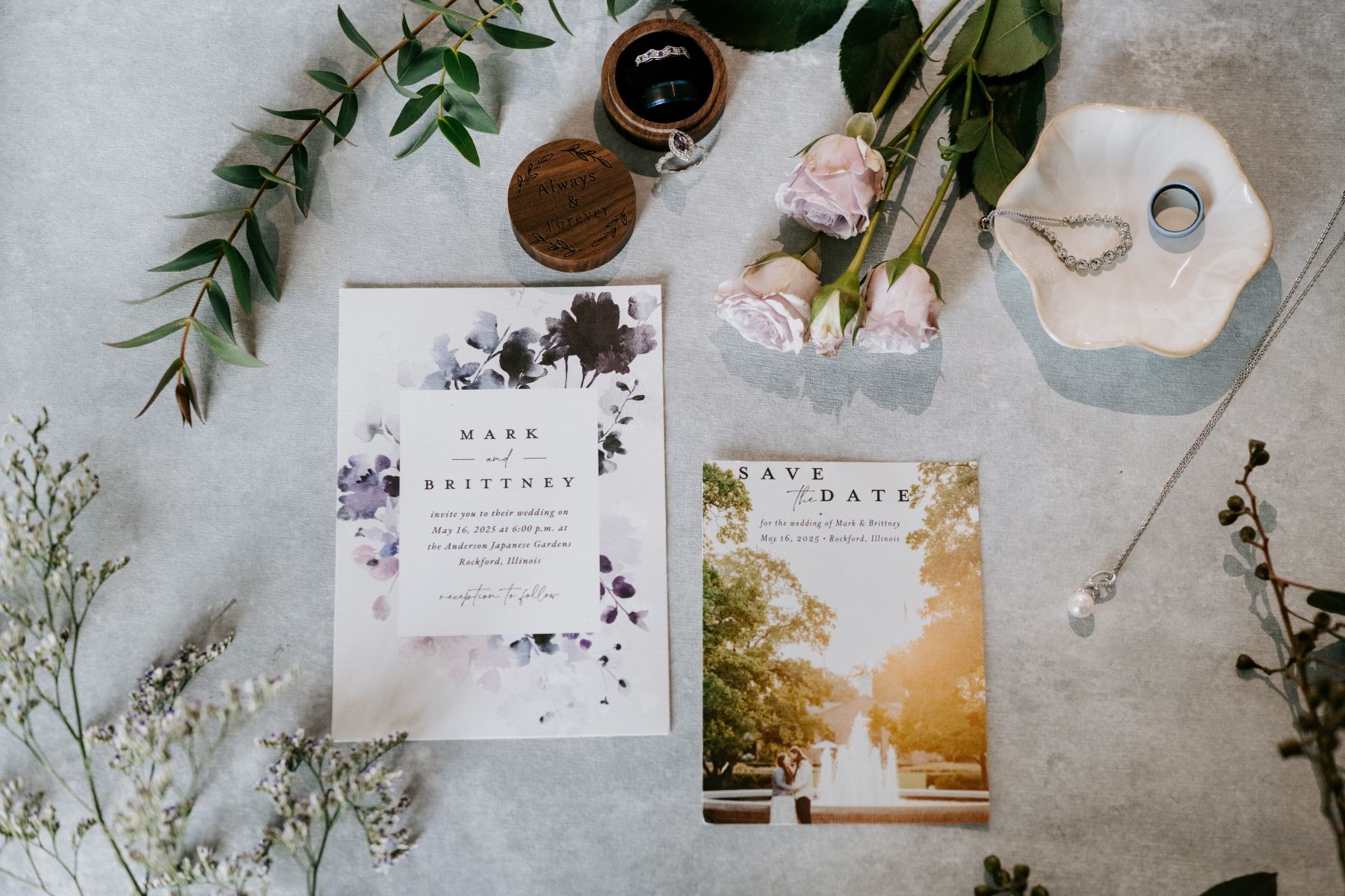 wedding details in a flat lay on a table including invitations, rings, and a necklace