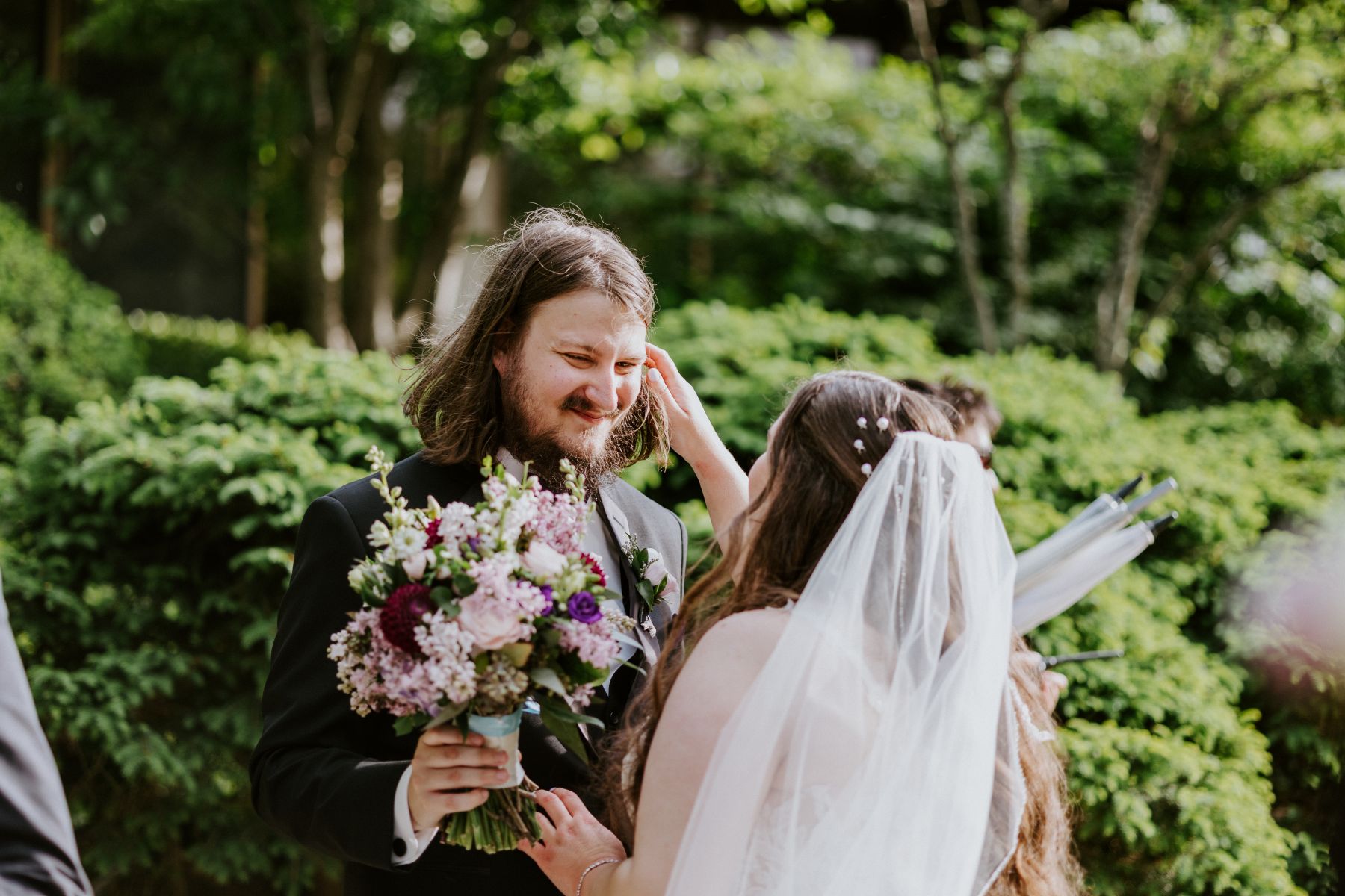 a woman wearing a white wedding dress and veil tucking a piece of hair behind her partner's ear he is wearing a black suit and holding flowers and smiling at her
