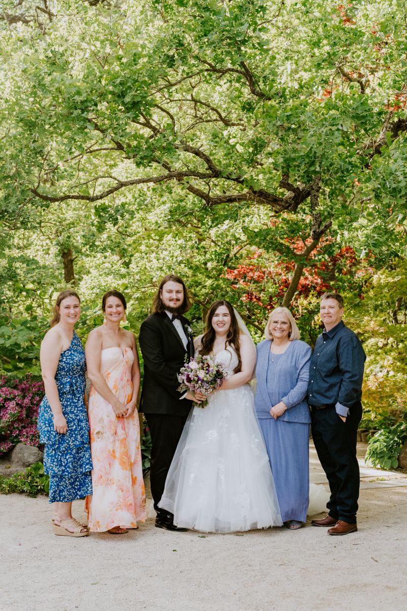 a couple posing with their families on the wedding they are all smiling and behind them is a variety of trees